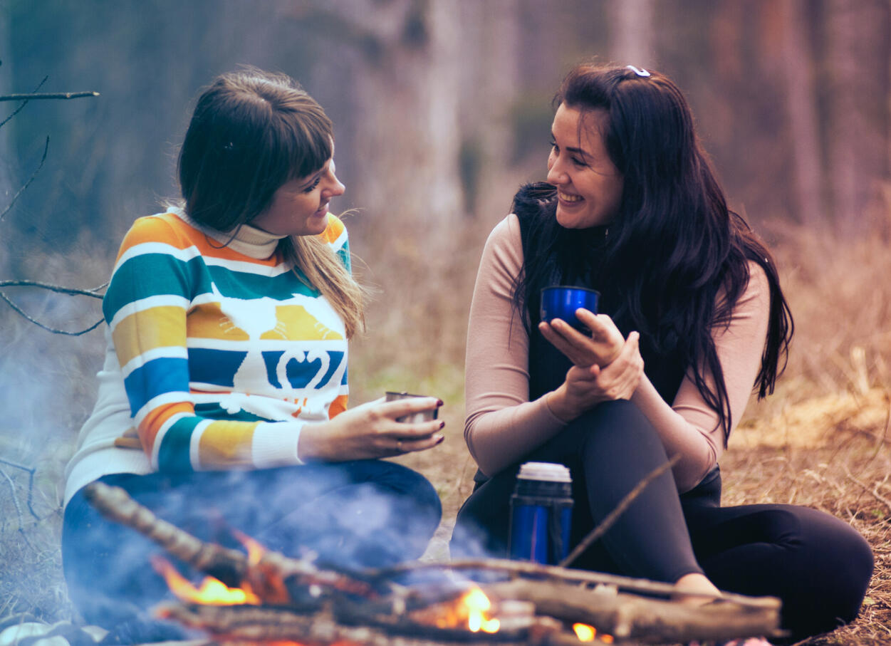 Woman on left in a yellow and green sweater looking at a woman in a white longsleeve shirt and vest. Both are holding a cup behind a camp fire in the woods.