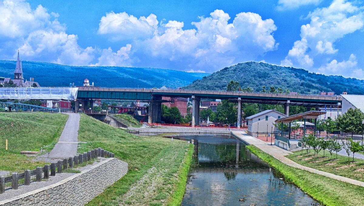 Canal end with a bridge behind and the mountains and city of Cumberland behind the bridge. The rolling hills of the Allegheny Mountains are green.